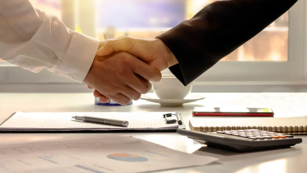 Deux bras qui se serrent la mains au premier plan, un contrat au second plan illustrant l'aspect contractuel d'un apporteur d'affaires et d'un agent commercial